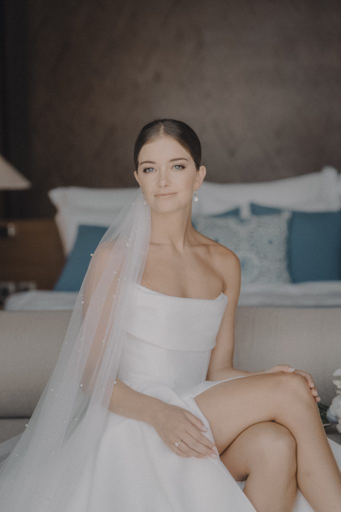 Bride in a white gown and veil sitting on a villa bed